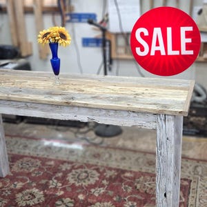 May include: A rustic wooden table with a distressed finish, featuring a light gray base and a natural wood top. A blue vase with yellow flowers sits on the table. A red circle with the word "SALE" is in the background.