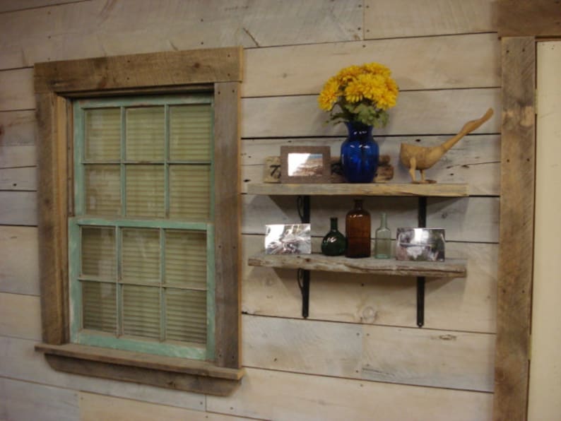 May include: Rustic wooden shelves with a blue glass vase filled with yellow flowers, a wooden bird figurine, and various bottles. The shelves are mounted on a whitewashed wood wall with a window to the left.