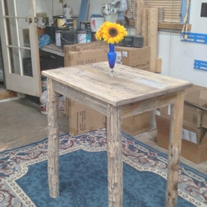 May include: A rustic, rectangular wooden table with a light brown finish. The table has four sturdy legs and a sunflower in a blue vase on top. The table is placed on a blue patterned rug.