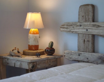 May include: A rustic wooden headboard with a white lamp with a cream shade on a wooden side table. A small potted cactus is on the table next to the lamp.