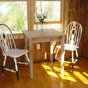 May include: A small, square wooden table with two white chairs. The chairs have black accents on the legs and back. A vase of white flowers sits on the table. The room has wood paneling and a window.