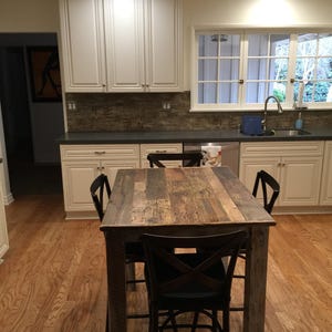 May include: A rustic wooden table with a distressed finish and four black chairs. The table is in a kitchen with white cabinets and a gray tiled backsplash.