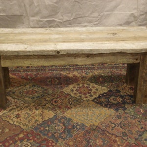 May include: A rustic wooden bench with a weathered gray finish. The bench has a simple design with four legs and a rectangular top. The bench is sitting on a patterned rug.