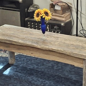 May include: A rectangular wooden bench with a weathered, light brown finish and four sturdy legs. A small, cobalt blue glass vase containing three yellow sunflowers with dark brown centers sits in the center of the bench top surface.