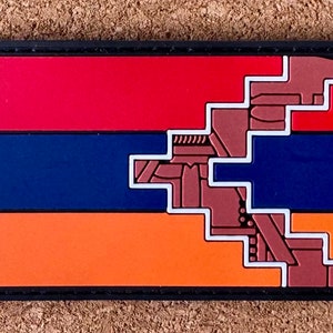 Puede incluir: Un parche rectangular con el diseño de la bandera armenia. La bandera está dividida en tres bandas horizontales de color rojo, azul y naranja, con un diseño geométrico marrón y blanco en el lado derecho. El parche tiene un borde negro.
