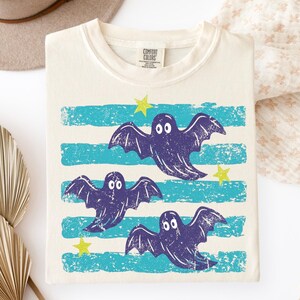 Puede incluir: Una camiseta color marfil con un estampado gráfico de tres murciélagos de color morado oscuro volando sobre rayas horizontales de color turquesa desgastado. Pequeñas estrellas amarillas están dispersas entre los murciélagos. Etiqueta Comfort Colors.
