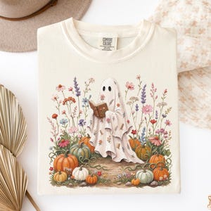 May include: An ivory t-shirt featuring a graphic of a ghost draped in a floral sheet, holding an open book. The ghost is surrounded by colorful wildflowers and various pumpkins in shades of orange, green, and white. The shirt has a crew neckline.
