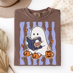 May include: A brown crewneck t-shirt featuring a graphic of a white ghost reading a book titled Spooky Reads. The design includes light blue vertical stripes, orange jack-o'-lanterns, falling autumn leaves, and a steaming orange mug.