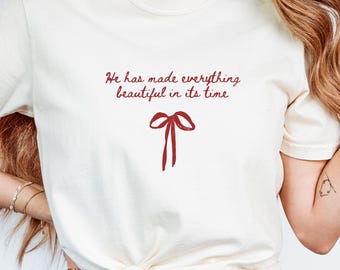 Può includere: T-shirt color avorio con la scritta "He has made everything beautiful in its time" e un fiocco rosso. La maglietta è annodata in vita e abbinata a jeans blu. La maglietta è a maniche corte e di stile casual.