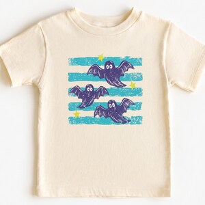 Puede incluir: Una camiseta de manga corta color crema con un estampado gráfico de tres fantasmas de color morado oscuro volando sobre rayas horizontales de color turquesa desgastado, acentuado por pequeñas estrellas amarillas texturizadas.
