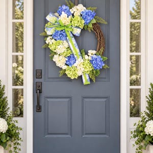 May include: A round wreath featuring a brown twig base decorated with blue, white, and light green hydrangea blossoms. The arrangement includes green foliage and a decorative bow made of blue and white patterned ribbon with lime green trim.