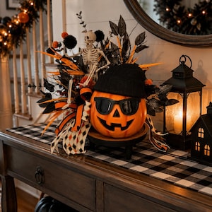 May include: A carved orange pumpkin wearing black sunglasses and a black beanie, positioned on a small wooden stand. The pumpkin is surrounded by black and orange foliage, a small skeleton figure, and patterned ribbons, creating a festive display.