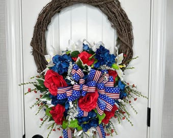 May include: A patriotic wreath with red, white, and blue flowers and a bow with stars and stripes. The wreath is made of twigs and is hanging on a white door.