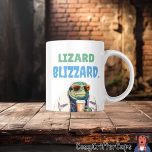 May include: White ceramic mug with a green lizard wearing sunglasses and a fur coat holding a cup of coffee. The mug has the text "Lizard Blizzard." printed on it.