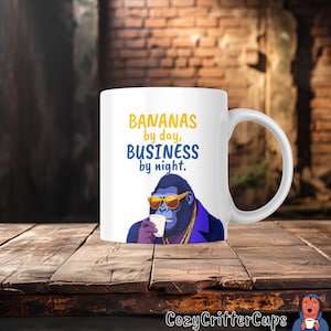 May include: White ceramic mug with a blue gorilla wearing sunglasses and a gold chain, drinking from a white cup. The text on the mug reads "BANANAS by day, BUSINESS by night."