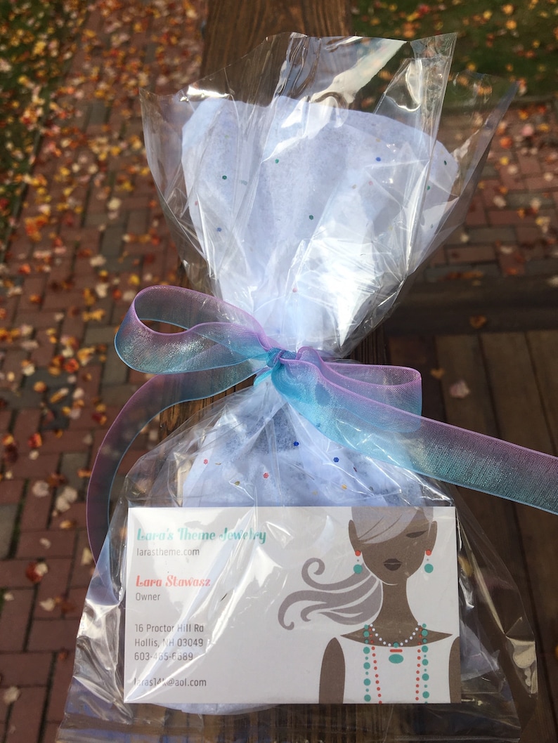 May include: A white gift bag with a blue and pink ribbon, containing a business card for Lara's Theme Jewelry. The business card features a woman with long hair wearing a turquoise necklace and earrings. The text on the card reads "Lara's Theme Jewelry", "larastheme.com", "Lara Stawasz", "Owner", "16 Proctor Hill Rd", "Hollis, NH 03049", "603-465-5389", and "ras@aol.com".