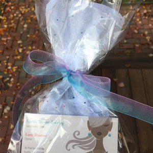 Op de afbeelding: Een cadeau verpakt in doorzichtig cellofaan, vastgebonden met een doorschijnend blauw en paars lint. Het pakket bevat een visitekaartje voor Lara's Theme Jewelry, met een gestileerde illustratie van een vrouw die sieraden draagt.