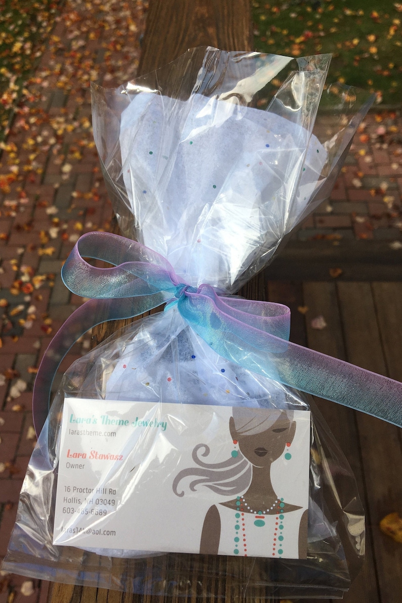 Op de afbeelding: Een cadeau verpakt in doorzichtig cellofaan, vastgebonden met een doorschijnend blauw en paars lint. Het pakket bevat een visitekaartje voor Lara's Theme Jewelry, met een gestileerde illustratie van een vrouw die sieraden draagt.