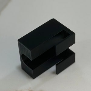 Puede incluir: Un objeto de plástico negro impreso en 3D con forma rectangular y un recorte en un lado. El objeto tiene una superficie lisa y parece ser parte de un ensamblaje más grande.