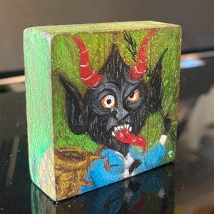 Peut inclure: Un petit bloc de bois peint à la main représentant une figure démoniaque. La créature a une fourrure noire, des cornes rouges et une longue langue rouge. Il est placé sur un fond vert avec une texture boisée. La figure porte une chemise bleue.