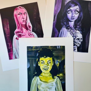 May include: Three framed illustrations of women with pale skin and dark hair. The women are depicted in a gothic style with dramatic makeup and clothing. The illustrations are set against dark backgrounds.