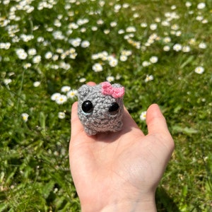 May include: A small grey crocheted animal with black eyes and a pink bow. The animal is being held in a hand against a background of green grass and white flowers.