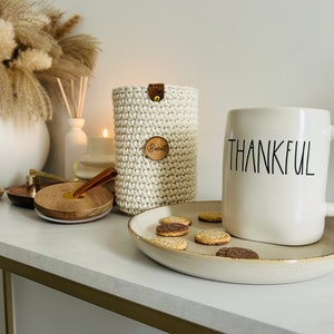 May include: White ceramic mug with the word "THANKFUL" in black lettering, a plate of biscuits, a crocheted white cup cosy with a wooden tag that says "Brewed", and a wooden coaster on a white countertop.