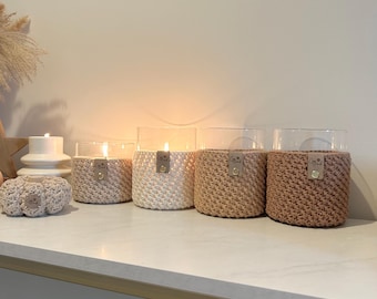 May include: A collection of glass candle holders with crochet sleeves in various neutral colors, including beige, cream, and brown. Each candle holder has a lit candle inside. A small crochet pumpkin is also visible.