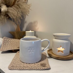 May include: A white ceramic mug with a snowflake design rests on a beige crocheted coaster. A lit candle is visible in a white ceramic oil diffuser with a star cutout. Decorative pampas grass and a wooden star are in the background.