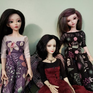 May include: Three dolls wearing different dresses. The doll on the left is wearing a white and purple dress with floral patterns. The doll in the middle is wearing a black and red dress. The doll on the right is wearing a black and white dress with floral patterns.