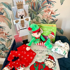 May include: A festive assortment of Christmas items. The image features a plush elf, children's clothing, socks, and decorative boxes with nutcracker designs. The clothing includes a grey t-shirt with an elf graphic and the text "That's not my elf..." and a white onesie.