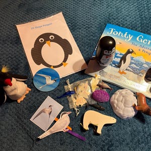 May include: A selection of penguin-themed items, including a plush toy, wooden figures, a book titled "Tonty Gentoo", and a 2D shape penguin card. Various other animal figures are also present, all on a blue surface.