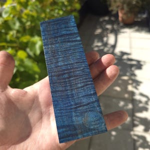 May include: A rectangular piece of wood with a vibrant blue finish and dark grain patterns. The wood is held in a hand, showcasing its polished surface and unique color variations. The wood appears to be approximately 5 inches long.