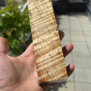 Könnte beinhalten: Ein rechteckiger Block aus gemasertem Holz, gehalten in einer Hand. Das Holz hat eine hellbraune Grundfarbe mit dunkleren, braunen, wellenförmigen Maserungen und feinen Linien. Das Holz scheint frisch geschnitten und für Holzbearbeitungsprojekte bereit zu sein.