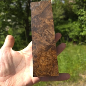 Könnte beinhalten: Ein rechteckiges Holzstück mit einem wirbelnden, natürlichen Maserungsmuster in Braun- und Beigetönen. Das Holz hat eine polierte, glänzende Oberfläche. Es wird in einer Hand vor einem Hintergrund aus grünem Laub gehalten.