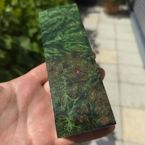 May include: A rectangular piece of wood with a vibrant green and brown burl grain pattern. The wood has a glossy finish, and the colours blend together to create a unique, natural design. The wood is held in a hand.