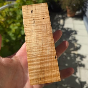 Könnte beinhalten: Ein rechteckiges Stück Ahorn-Holz mit Maserung, das eine warme, hellbraune Farbe mit wellenförmigen Maserungsmustern aufweist. Das Holz hat kleine, dunkle Knoten und Unvollkommenheiten, die seinen natürlichen Charakter unterstreichen. Das Holz wird in einer Hand gehalten.