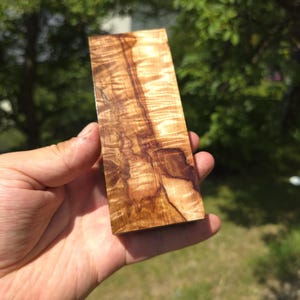 Könnte beinhalten: Ein rechteckiger Block aus poliertem Holz mit einer markanten Maserung, die durch wirbelnde hellbraune und dunkelbraune Streifen gekennzeichnet ist. Die Oberfläche hat ein glattes, reflektierendes Finish, das die natürliche Textur betont.