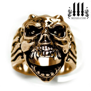 Puede incluir: Un anillo de calavera de color bronce con detalles, incluyendo dientes afilados y cuencas de los ojos. El anillo tiene una superficie texturizada y una banda ancha. El sitio web 3rexes.com es visible en el fondo.