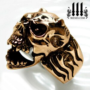 Puede incluir: Un anillo de calavera dorado con un diseño detallado. La calavera tiene la boca abierta con dientes y una superficie texturizada. La banda del anillo tiene un patrón ondulado.