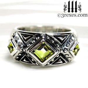 May include: A silver ring with a geometric design and three green gemstones. The ring has a textured surface and a wide band.