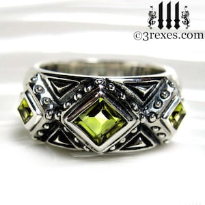 May include: A silver ring with a geometric design and three green gemstones. The ring has a textured surface and a wide band.