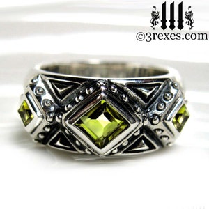 May include: A silver ring with a geometric design and three green gemstones. The ring has a textured surface and a polished finish.