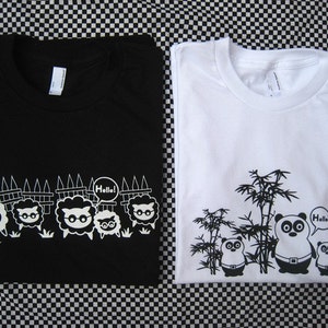 Può includere: Due t-shirt con design diversi. La t-shirt nera ha una fila di pecore con gli occhiali e la t-shirt bianca ha un panda con il bambù e il testo "Hello!"