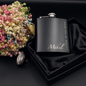May include: A black hip flask engraved with "FATHER OF THE BRIDE" and the name "Mikel" in a gift box with black satin lining. A bouquet of colourful flowers is next to the flask.