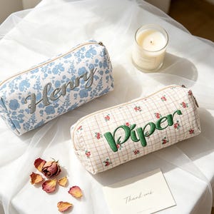 May include: Two personalized pencil cases. One is light blue with a floral pattern and the name "Henry" embroidered in gray. The other is beige with a plaid pattern and red floral accents, with the name "Piper" embroidered in green.