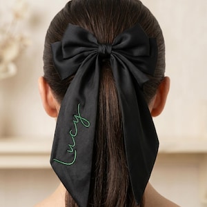 May include: A black satin hair bow with long streamers. The word "Lucy" is embroidered in green script on one streamer. The bow is tied at the back of the head, with the person's hair pulled back into a ponytail.