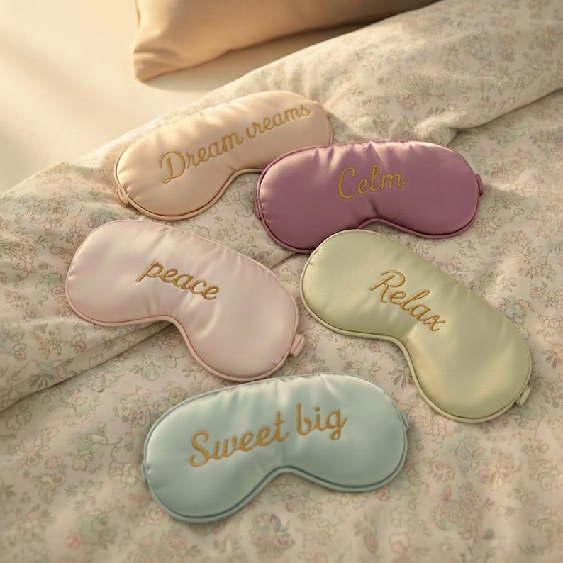 May include: Five satin sleep masks in pastel colors: peach, lavender, pink, mint green, and light blue. Each mask has gold embroidered text: "Dream dreams", "Calm", "peace", "Relax", and "Sweet big". The masks are arranged on a floral patterned bedspread.