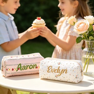 May include: Two personalized pencil cases on a table. One is pink with "Aaron" embroidered in green, the other is white with floral patterns and "Evelyn" in gold. A cupcake is held between two children.