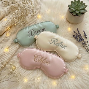 May include: Three satin sleep masks in pastel colors: mint green with "Dream" embroidered, cream with "Relax", and pink with "Chloe". The masks are on a fluffy white surface, with fairy lights, a succulent, and lavender sprigs.